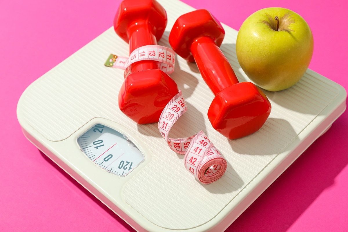 weigh scales, apple, dumbbells and measuring tape on pink backgr